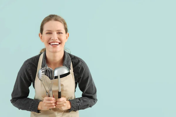 Mooie jonge vrouw in schort en met keukengerei op kleur achtergrond — Stockfoto