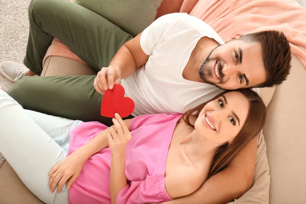 Retrato de feliz pareja joven con corazón de papel sentado en el sofá en casa — Foto de Stock