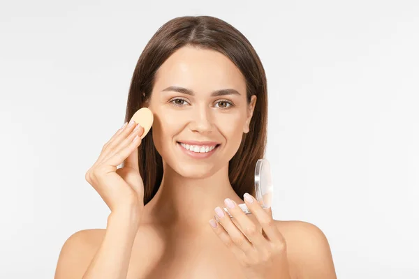 Beautiful young woman applying facial powder against light background — Stock Photo, Image