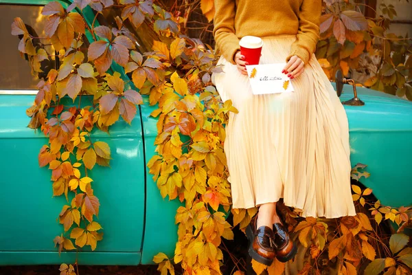 Schöne junge Frau mit Tasse Kaffee und Grußkarte sitzt an einem Herbsttag im Freien auf dem Auto — Stockfoto