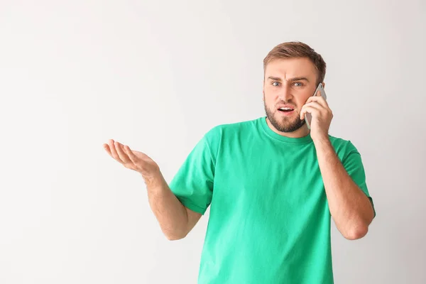 Confused young man talking by phone on light background — Stock Photo, Image