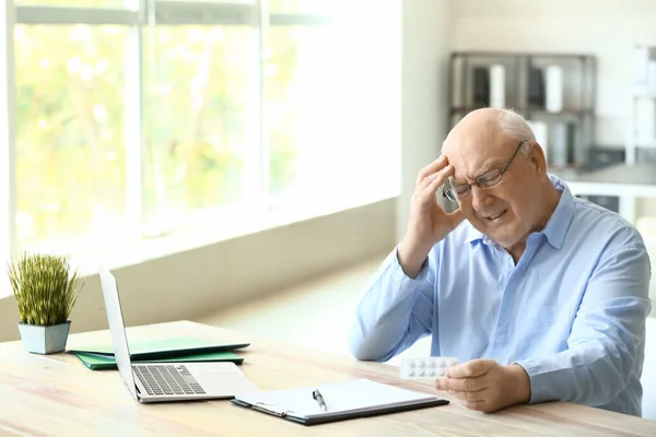 Elderly man with pills suffering from headache in office — Stock Photo, Image