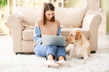 Evde dizüstü bilgisayar kullanan sevimli köpekli güzel genç bir kadın.