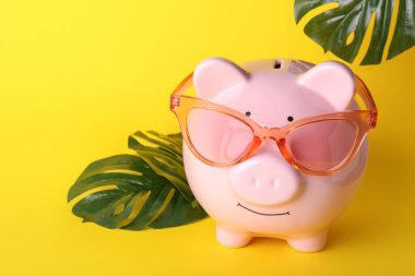 Piggy bank with sunglasses and tropical leaves on color background clipart