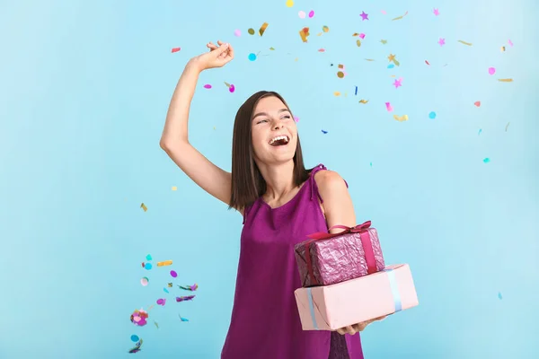 Schöne junge Frau mit Geschenken und fallendem Konfetti auf farbigem Hintergrund — Stockfoto