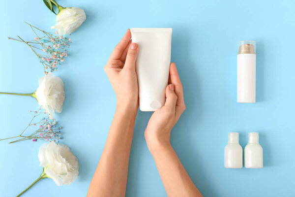 Hands of young woman, different creams and flowers on color background