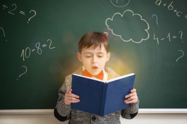 Sınıfta tahtanın yanında kitabı olan küçük bir okul çocuğu.