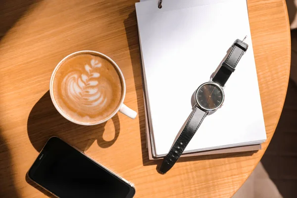 Cup of tasty coffee with notebook, clock and mobile phone on table in cafe