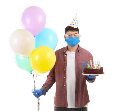 Beyaz arka planda parti dekoru ve doğum günü pastası olan koruyucu maskeli bir Asyalı. Salgın kavramı