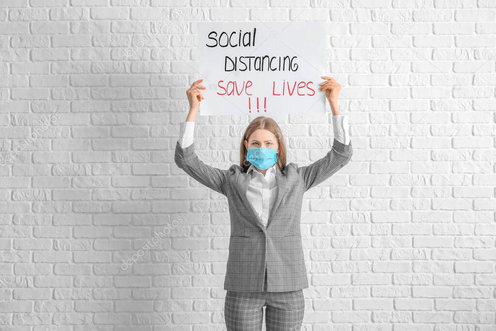 Young woman holding paper with text SOCIAL DISTANCING. SAVE LIVES on white brick background. Concept of epidemic