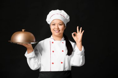 Tepsisi olan güzel bir Asyalı aşçı koyu arka planda iyi niyet gösterisi yapıyor.