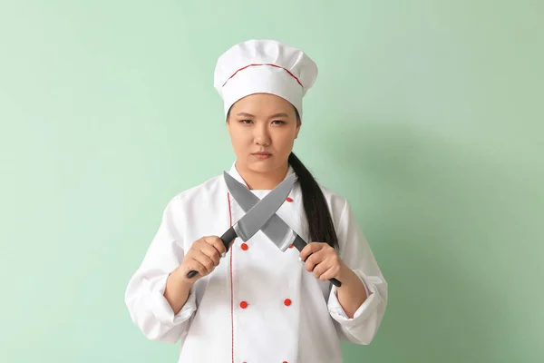 Beautiful Asian chef with knives on color background
