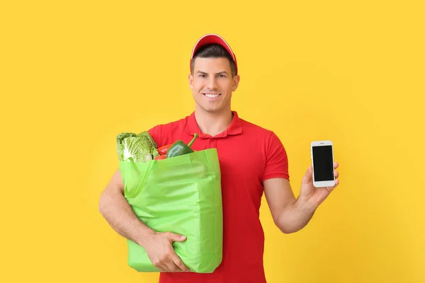 Delivery man with food in bag and mobile phone on color background