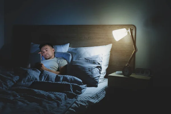 Young man suffering from sleep deprivation with tablet computer in bedroom at night