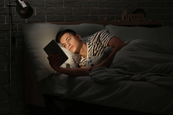 Young man suffering from sleep deprivation with tablet computer in bedroom at night