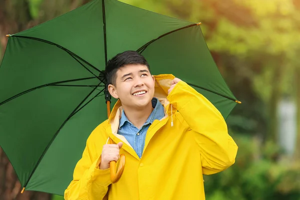 Young Asian man with umbrella wearing raincoat in park