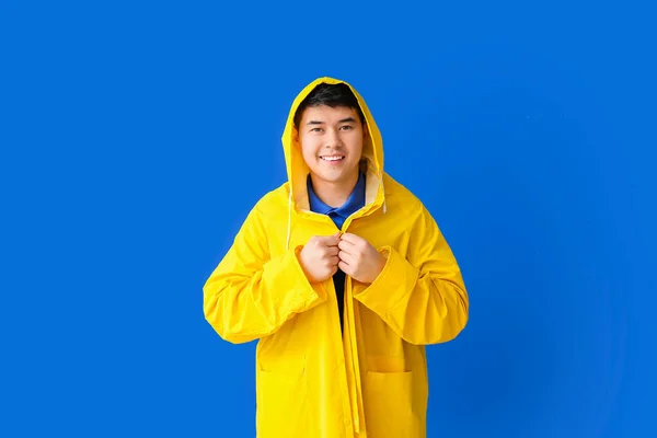 Young Asian man in raincoat on color background