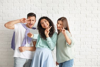 Genç arkadaşlar beyaz tuğla arka planda selfie çekiyorlar.