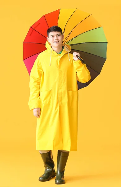 Young Asian man in raincoat and with umbrella on color background