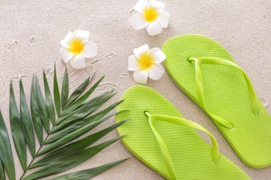 Havalı parmak arası terlikler, çiçekler ve kumdaki tropikal yapraklar.