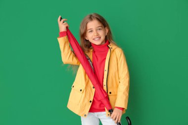 Yağmurluk giymiş şirin bir kız ve arka planda şemsiyesi var.