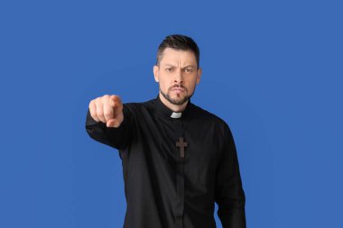 Angry priest pointing at viewer on color background clipart