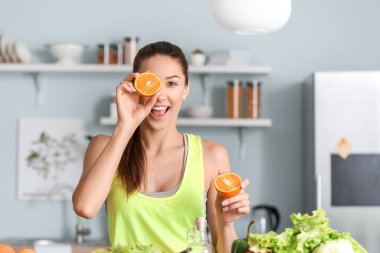 Beautiful young woman with fresh orange fruit in kitchen clipart