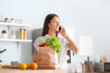 Marketten taze ürünleri olan genç bir kadın evde cep telefonuyla konuşuyor.