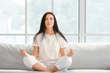 Evdeki kanepede meditasyon yapan genç bir kadın.