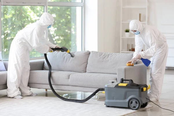 Workers in biohazard costume removing dirt from sofa in house