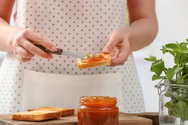 Woman spreading tasty peach jam on bread slice in kitchen