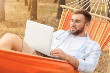 Bilgisayarlı genç adam dışarıda hamakta dinleniyor.