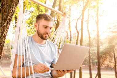 Bilgisayarlı genç adam dışarıda hamakta dinleniyor.