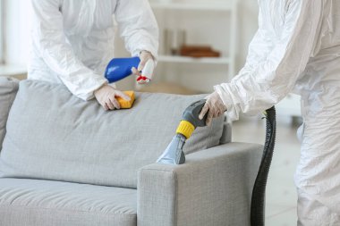 Biyolojik tehlike kostümlü işçiler evdeki kanepedeki kiri temizliyorlar.