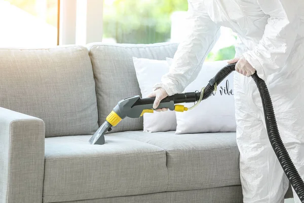 Worker in biohazard costume removing dirt from sofa in house