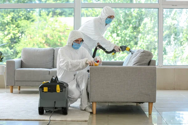 Workers in biohazard costume removing dirt from sofa in house