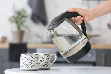 Kadın mutfak masasındaki bardağa elektrikli su dolduruyor.