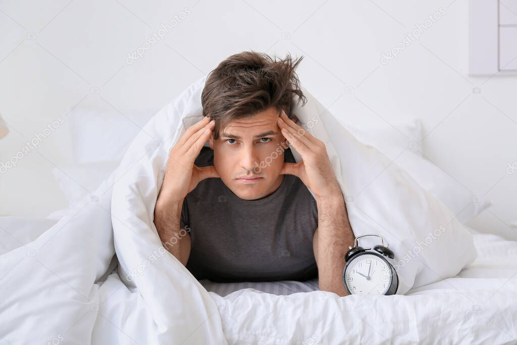 Young man with alarm clock suffering from insomnia in bedroom