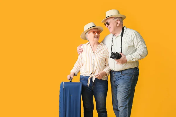 Happy senior couple with packed suitcase on color background