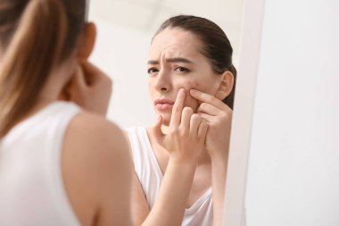 Young woman with acne problem squishing pimples near mirror clipart