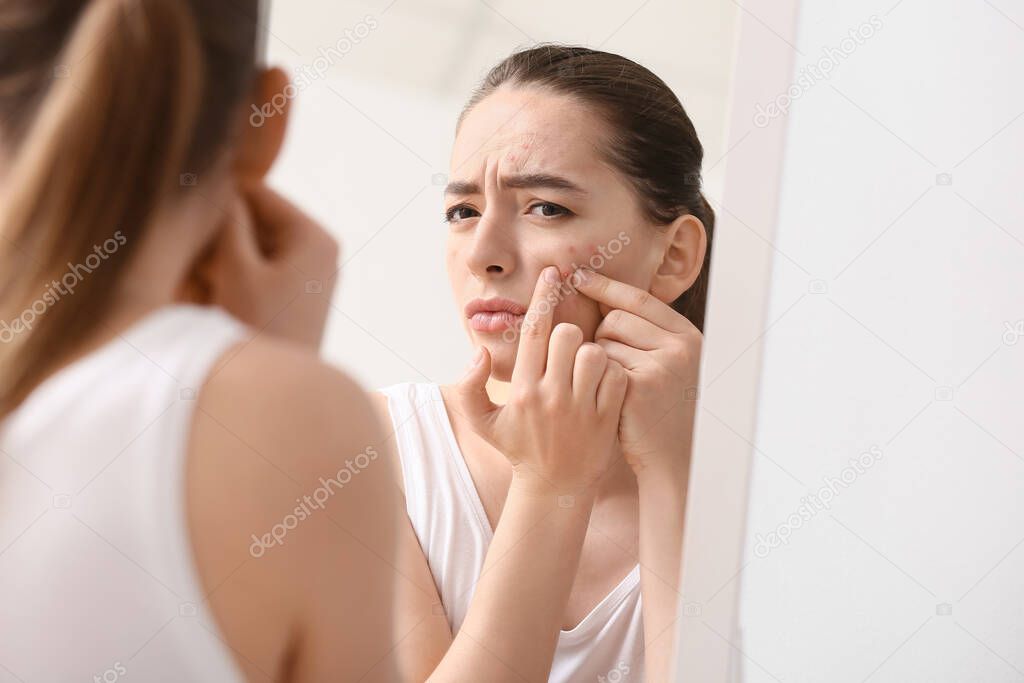 Young woman with acne problem squishing pimples near mirror