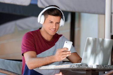 Kulaklıklı ve cep telefonlu bir adam açık kafede dizüstü bilgisayarla çalışıyor.