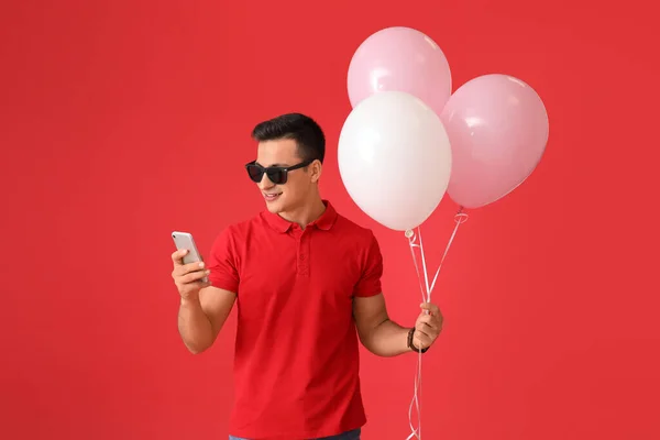 Young man with balloons and mobile phone on color background