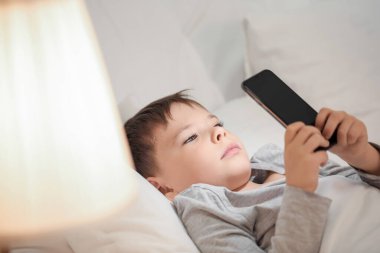 Küçük çocuk yatak odasında cep telefonuyla uykusuzluk çekiyor.