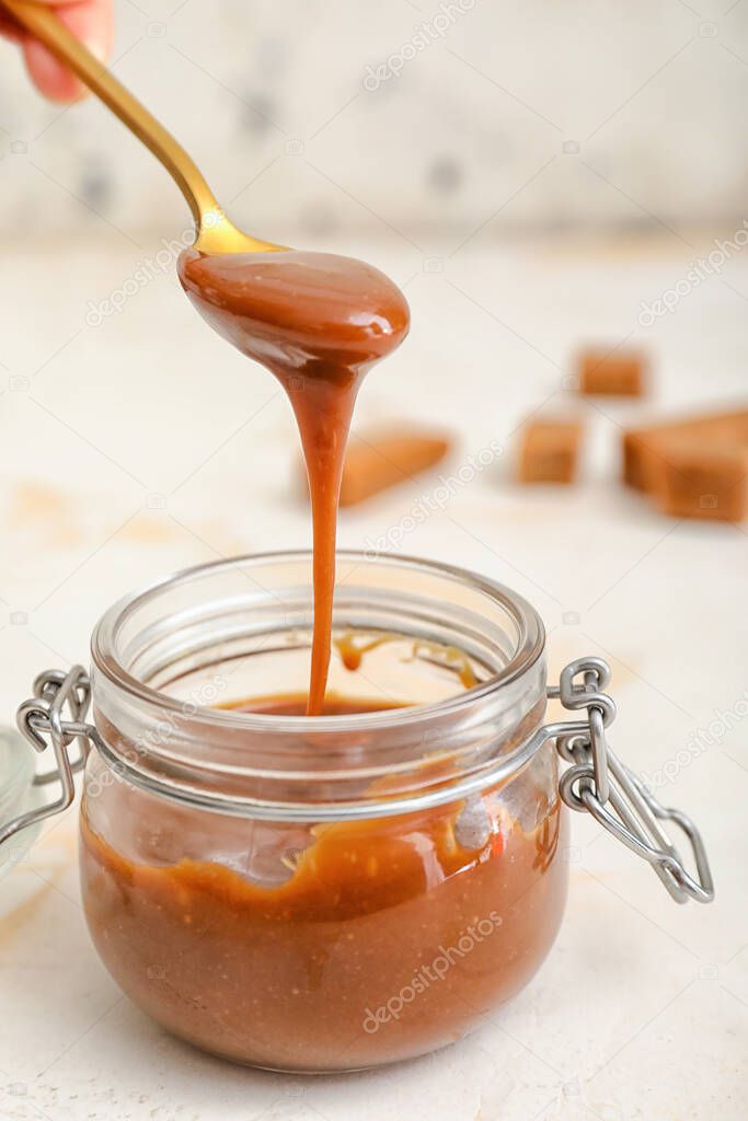 Jar and spoon with liquid caramel on white background