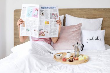 Genç bir kadın gazete okuyor ve yatakta lezzetli bir kahvaltı yapıyor.