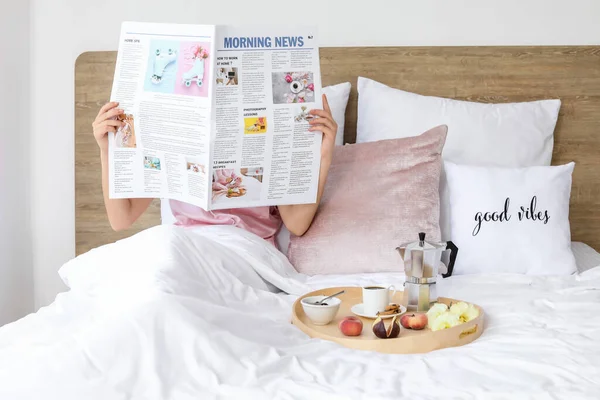 Genç bir kadın gazete okuyor ve yatakta lezzetli bir kahvaltı yapıyor.