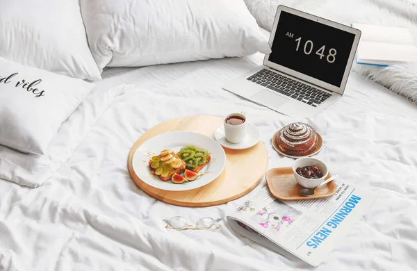 Tray with tasty breakfast, laptop and newspaper on bed