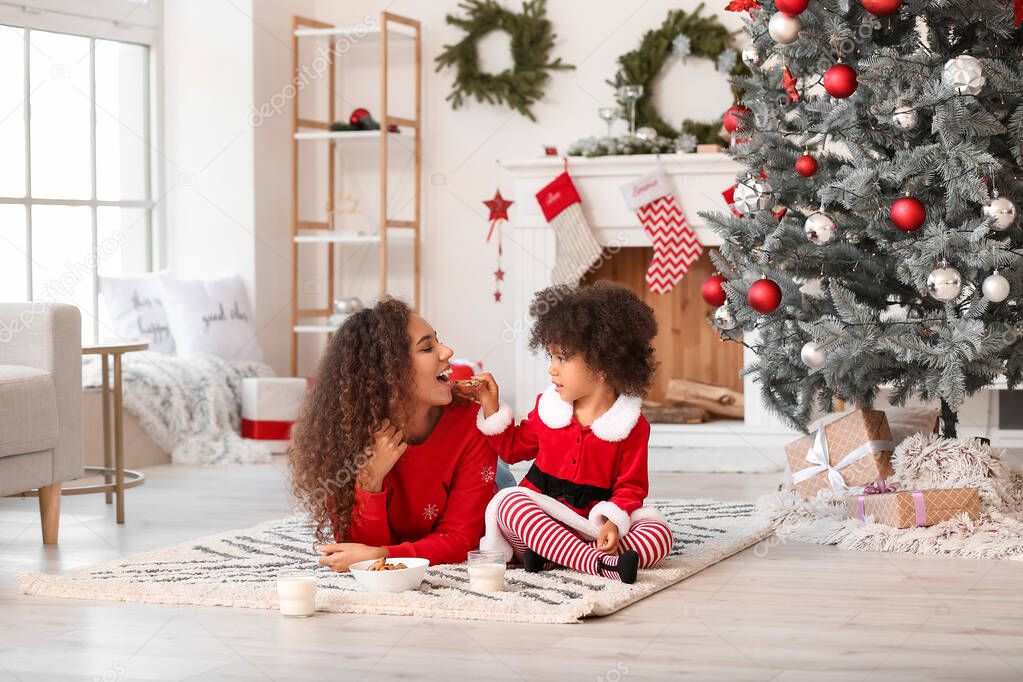 Cute African-American girl with her mother eating cookies and drinking milk at home on Christmas eve