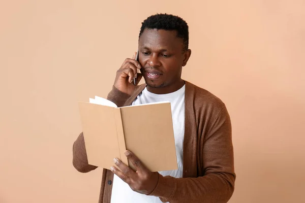 African-American man with book talking by phone on color background